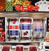A close-up view of a slot machine with vibrant colors and various gambling symbols such as dice, coins, and playing cards. The reels display a combination of symbols, including bars, cherries, and the number seven. The chrome exterior and colorful details convey the theme of casino and gambling.