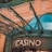 The image displays the entrance to a casino, characterized by detailed and ornate architectural elements with green metal structures and a sign that reads 'CASINO'. The background includes a glass dome and a peach-colored wall.