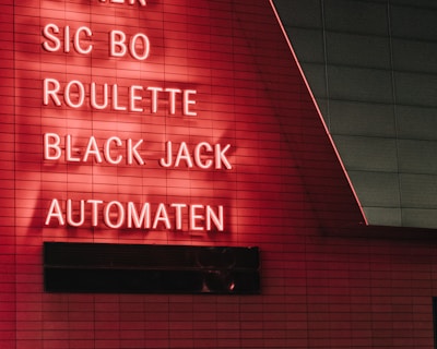 Red neon letters are mounted on a dark tiled wall, displaying the words Poker, Sic Bo, Roulette, Black Jack, and Automaten.