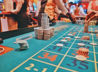 A casino scene with a roulette table features prominently, covered in green felt with colorful numbers and betting squares. Several stacks of poker chips are placed on different numbers, and the hands of several people are visible as they interact with the table. In the background, there are slot machines and more people, creating a vibrant and busy atmosphere.