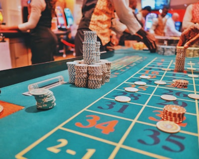 A casino scene with a roulette table features prominently, covered in green felt with colorful numbers and betting squares. Several stacks of poker chips are placed on different numbers, and the hands of several people are visible as they interact with the table. In the background, there are slot machines and more people, creating a vibrant and busy atmosphere.