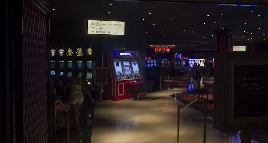 A dimly lit casino with several rows of slot machines, some displaying bright graphics and lights. There are a few people scattered throughout the area, engaging with the machines. The floor is reflective and polished, adding to the ambiance, while the ceiling is dotted with numerous small lights.