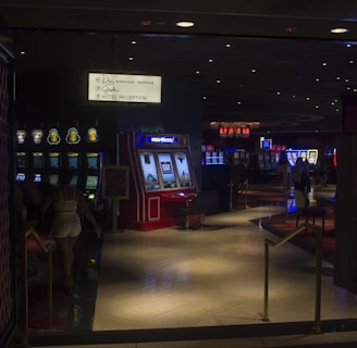 A dimly lit casino with several rows of slot machines, some displaying bright graphics and lights. There are a few people scattered throughout the area, engaging with the machines. The floor is reflective and polished, adding to the ambiance, while the ceiling is dotted with numerous small lights.