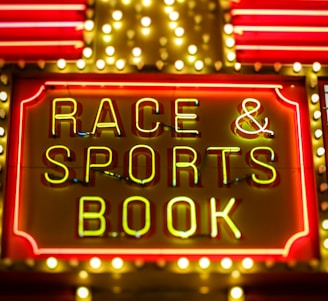 Bright neon lights on a marquee sign display the words 'RACE & SPORTS BOOK'. The sign is surrounded by rows of yellow and white lights against a red background, evoking a vibrant, energetic atmosphere typically associated with casinos or entertainment venues.