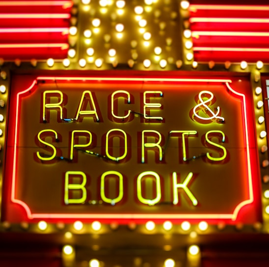 Bright neon lights on a marquee sign display the words 'RACE & SPORTS BOOK'. The sign is surrounded by rows of yellow and white lights against a red background, evoking a vibrant, energetic atmosphere typically associated with casinos or entertainment venues.