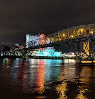 A brightly lit casino with neon signs and red lights is reflected in a river. A bridge stretches across the water, with an urban skyline visible in the background against an overcast night sky.