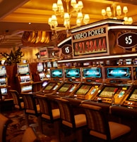 A casino floor featuring rows of brightly lit video poker machines. The area is illuminated by elegant chandeliers, with a display board showing a large sum of money, suggesting a possible jackpot. The rich decor includes luxurious carpets and comfortable seating, adding to the opulent atmosphere.