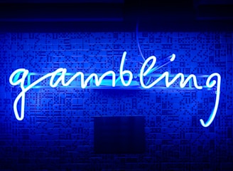 A bright blue neon sign spells out the word 'gambling' in cursive script. The background features a textured wall covered in domino tiles, also illuminated by the blue light, creating a vivid and visually striking scene.