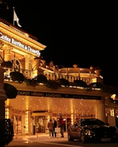 A luxurious casino at night, illuminated with warm, golden lights. The building features elegant architecture with columns and decorative elements. A few people are walking towards the entrance, and there are a couple of parked cars nearby.