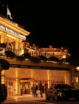 A luxurious casino at night, illuminated with warm, golden lights. The building features elegant architecture with columns and decorative elements. A few people are walking towards the entrance, and there are a couple of parked cars nearby.