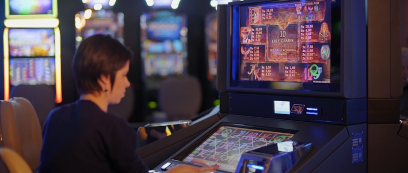 A person is seated at a slot machine console in a casino, focusing intently on the game displayed on the screen. The background shows several other illuminated gaming machines, adding a vibrant and colorful atmosphere to the scene.