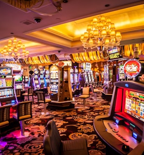 A vibrant casino interior filled with rows of colorful slot machines, each illuminated with bright lights and displays. The ceiling is adorned with elegant chandeliers and golden drapes, creating a luxurious atmosphere. The patterned carpet adds to the elaborate design of the space.