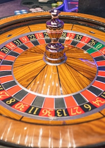 A roulette wheel with alternating red and black numbered pockets on a wooden platform. The wheel contains a small white ball resting in one of the pockets, indicating gameplay. Surrounding the wheel are stacks of colorful poker chips, suggesting a casino environment.