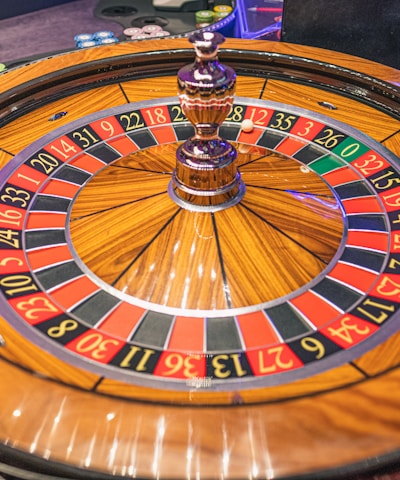 A roulette wheel with alternating red and black numbered pockets on a wooden platform. The wheel contains a small white ball resting in one of the pockets, indicating gameplay. Surrounding the wheel are stacks of colorful poker chips, suggesting a casino environment.