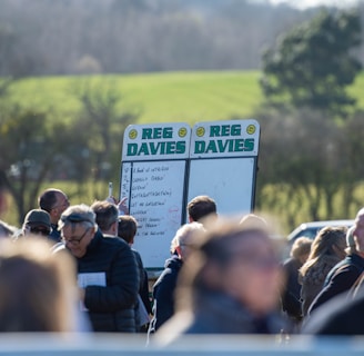 A group of people gathered outdoors with two large betting boards in the background labeled 'REG DAVIES'. The setting appears to be a countryside location with trees and fields visible in the distance. The crowd consists of individuals dressed in jackets and hats, engaging in conversation.