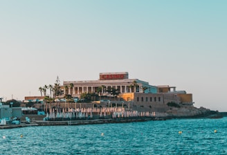 A seaside view of a large building labeled as a casino, situated on a rocky shore. The building is surrounded by palm trees and other smaller structures. The calm sea water reflects the sky, and there are several closed umbrellas lined along the waterfront.
