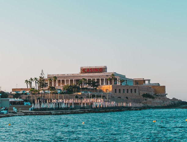 A seaside view of a large building labeled as a casino, situated on a rocky shore. The building is surrounded by palm trees and other smaller structures. The calm sea water reflects the sky, and there are several closed umbrellas lined along the waterfront.