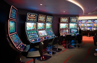 A row of brightly lit slot machines in a casino setting, each with colorful displays and various game themes. The machines are lined up against a dark wall and are unoccupied, with cushioned chairs placed in front of them. The area is dimly lit except for the glow from the screens, creating a vibrant and engaging atmosphere.