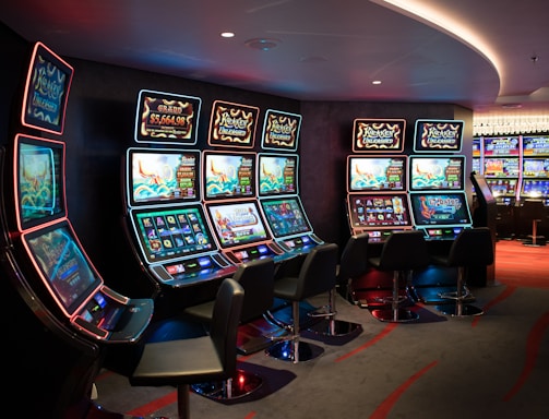 A row of brightly lit slot machines in a casino setting, each with colorful displays and various game themes. The machines are lined up against a dark wall and are unoccupied, with cushioned chairs placed in front of them. The area is dimly lit except for the glow from the screens, creating a vibrant and engaging atmosphere.