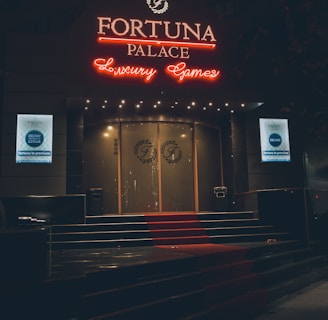 A neon-lit casino entrance with a red carpet leading up to large double doors. The sign reads 'Fortuna Palace Luxury Games' in bright red and white neon lights. The dimly lit atmosphere suggests a night-time setting. There are two digital displays on either side of the entrance showing promotional content.
