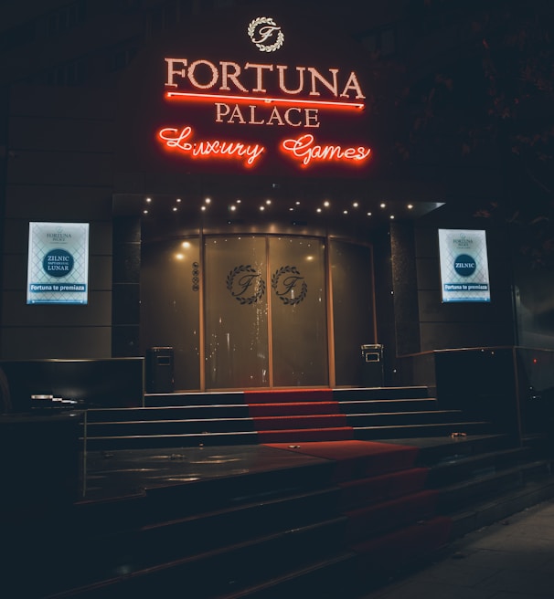 A neon-lit casino entrance with a red carpet leading up to large double doors. The sign reads 'Fortuna Palace Luxury Games' in bright red and white neon lights. The dimly lit atmosphere suggests a night-time setting. There are two digital displays on either side of the entrance showing promotional content.