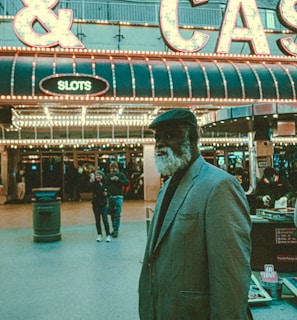 An older man with a gray beard and wearing a suit and hat stands in front of a brightly lit casino. The background features a large illuminated sign reading 'CASINO' and 'SLOTS' among other words, with numerous lights outlining the entrance. Several other people are visible in the background, engaged in various activities, some walking, some standing, and one looking at something on display.