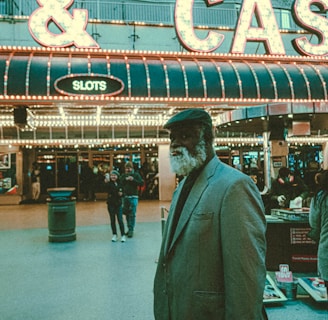 An older man with a gray beard and wearing a suit and hat stands in front of a brightly lit casino. The background features a large illuminated sign reading 'CASINO' and 'SLOTS' among other words, with numerous lights outlining the entrance. Several other people are visible in the background, engaged in various activities, some walking, some standing, and one looking at something on display.