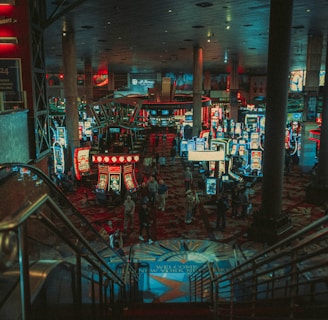A bustling casino floor filled with groups of slot machines and people walking around. The atmosphere is vibrant with neon lights and illuminated signs. The interior has a ceiling supported by large pillars and there are stairs leading down to the gaming area.