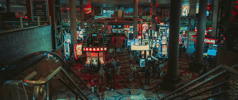 A bustling casino floor filled with groups of slot machines and people walking around. The atmosphere is vibrant with neon lights and illuminated signs. The interior has a ceiling supported by large pillars and there are stairs leading down to the gaming area.