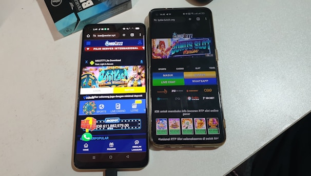 Two smartphones are displayed on a white surface, both showing gambling websites with colorful graphics, text, and various options for logging in and accessing games. The screens are bright and filled with promotional content related to casino and slot games.
